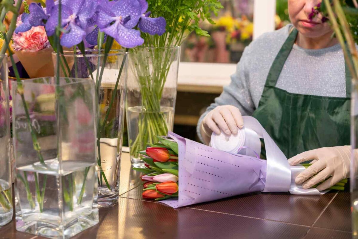 A woman wrapping a bunch of flowers and preserving some flowers in a transparent jar filled with water.