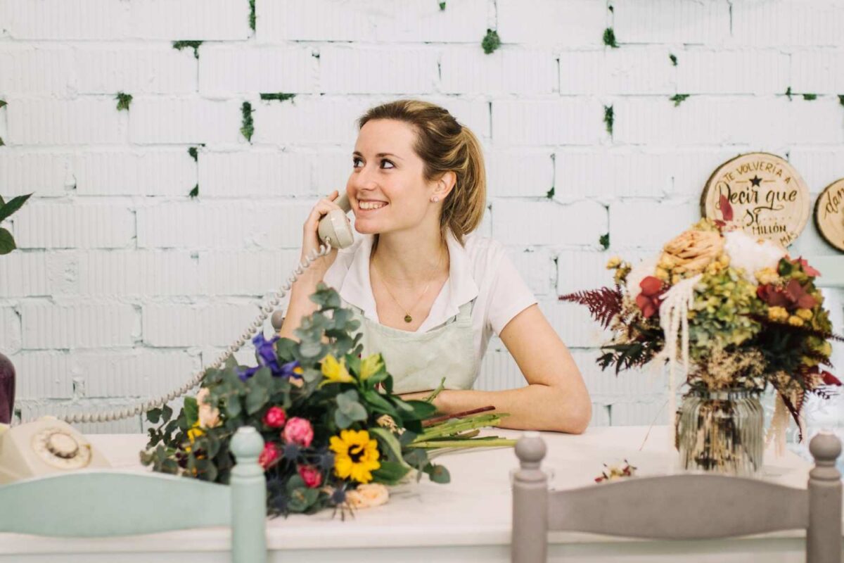 A cheerful young woman speaking on phone in a florist shop and taking flower order online.