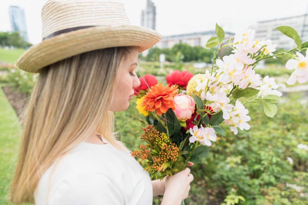 A young lady wearing a hat and holding a bunch of colorful flowers and smelling them