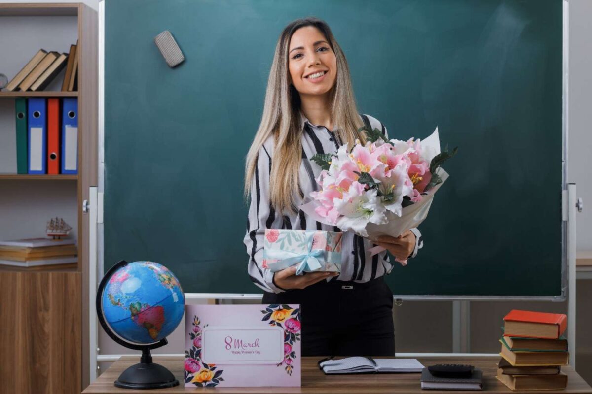 A young woman teacher standing near blackboard classroom holding flower bouquet present box from students.