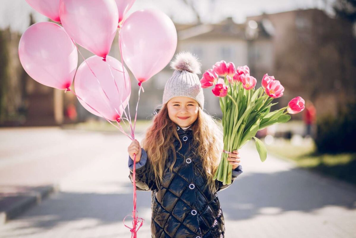 A little girl holding stems of flowers and some balloons.